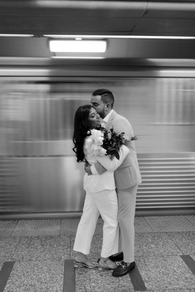 Woman and man in pantsuits stand in front of Chicago subway. Woman is holding a bouquet of roses while man kisses her on cheek