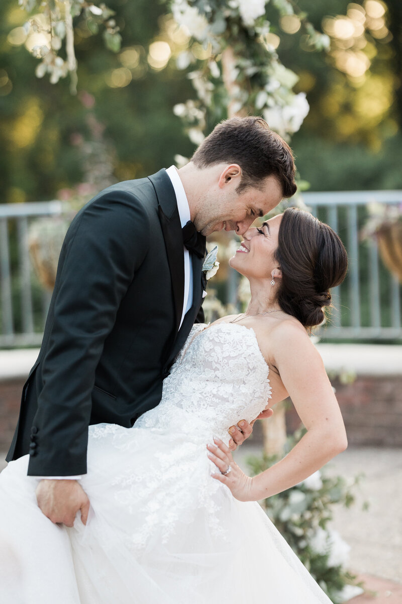 white groom in a black tux holding and dipping a white brunette bride in a strapless gown touching noses with eyes closed and smiling