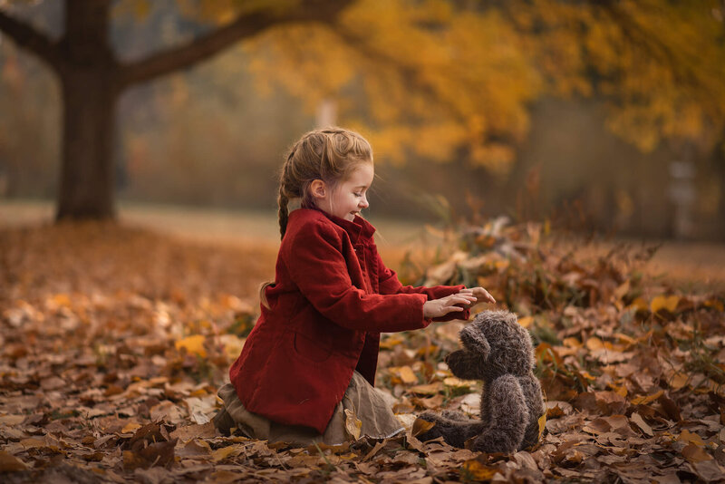 A Little girl in a red coat sits in a pile of fall leaves playing with a teddy bear. Captured in Kamloops by Brittany Danielle