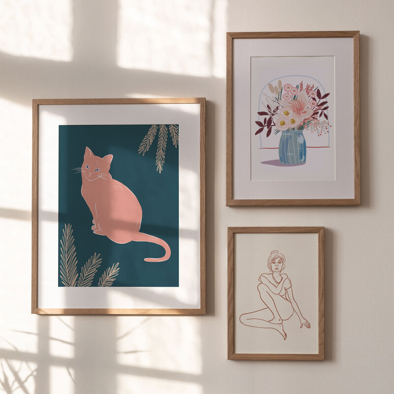 Three framed art prints by Skye McNeill hung on a sunlit wall in wooden frames. Featuring a cat, a bouquet of flowers and a line drawing of a figure. Advertising Skye's shop.