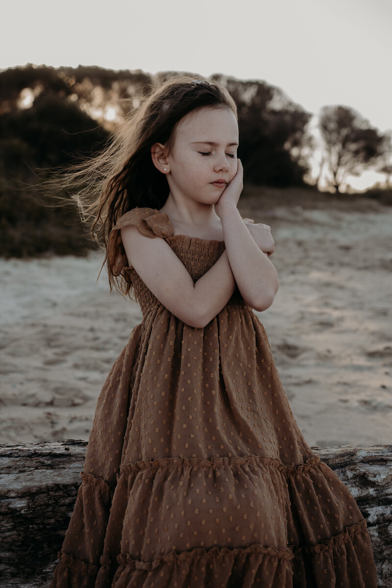 A young girl in a brown dress stands outdoors with her eyes closed and hand on her face, set against the serene backdrop of trees and a sandy area—an ideal moment captured by natural family photography NSW.