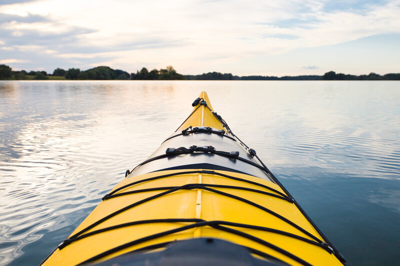 View from a yellow kayak on a calm lake, with rippling water and a distant tree-lined shore under a blue sky with scattered clouds.