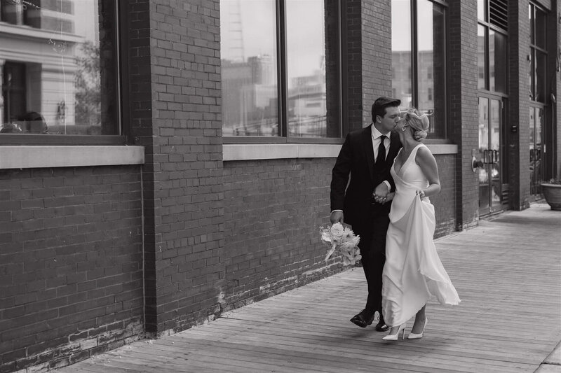 Groom holds bridal bouquet and bride holds dress as they kiss along a stroll.