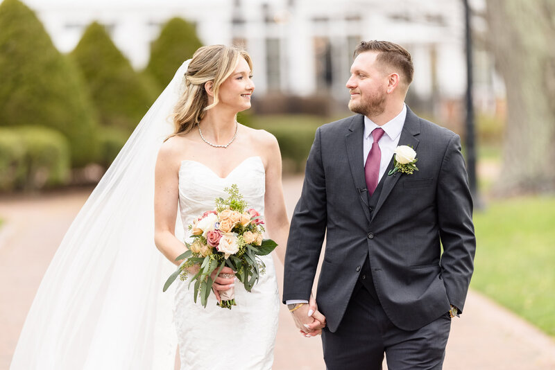 Bride and groom walking hand-in-hand outdoors, smiling lovingly at each other on their wedding day.