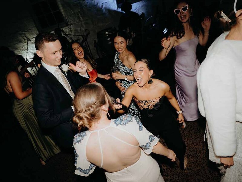 Wedding guests dancing and celebrating energetically on the dance floor during live acoustic performance.