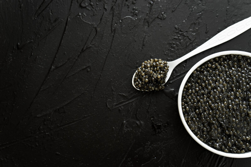 A white bowl of caviar on a black textured background with a spoon of caviar next to the bowl