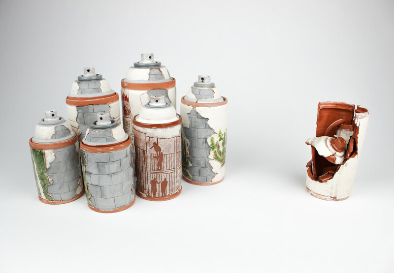 six spray paint can jars made out of ceramic are decorated with clay and underglaze to resemble stone blocks and broken walls with saguaro cacti , one jar sits to the side and is broken
