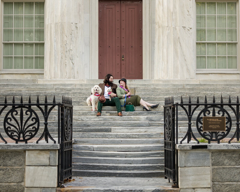 Engaged couple sitting on the steps of the Second Bank of the United States, enjoying the afternoon with their dogs during their Philly engagement session.