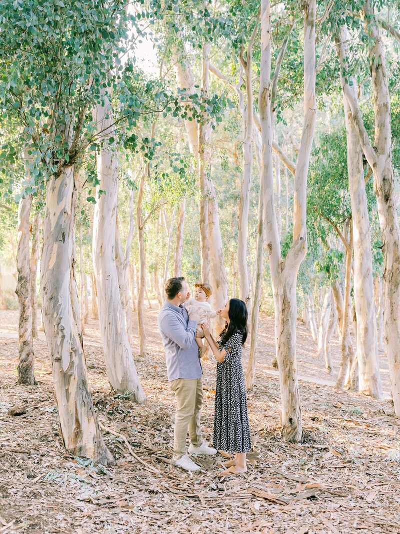 A couple joyfully holds a toddler between them in a sunlit eucalyptus forest, surrounded by tall, thin trees. The scene is serene and warm.