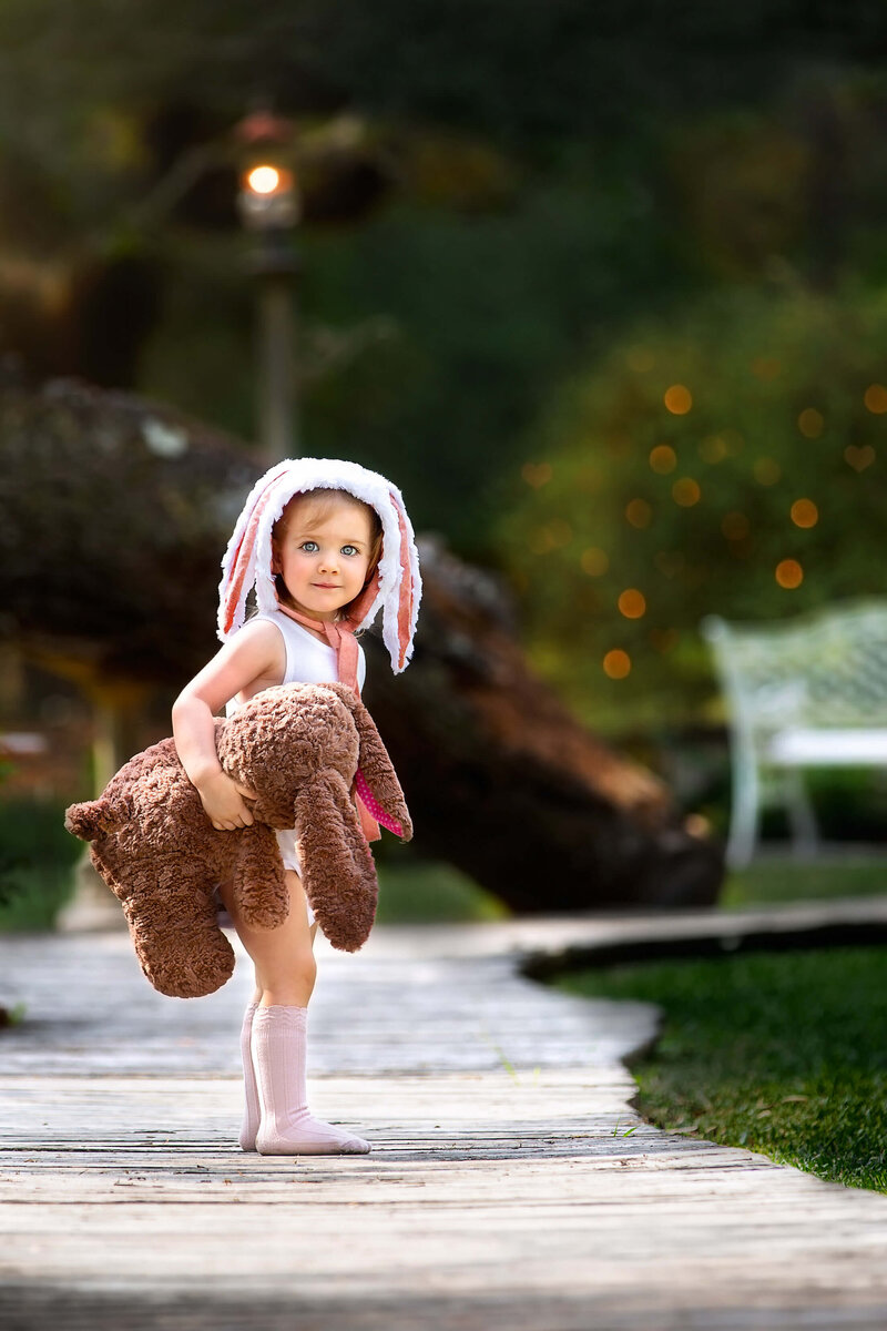 Adorable blue eyed toddler wearing a bunny bonnet while holding a stuffed bunny at  the beautiful location Out Under the Trees in Texas.