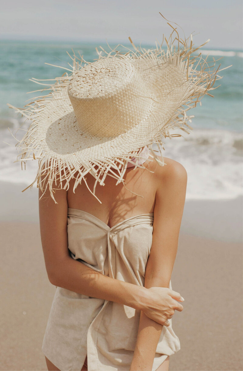 Woman traveler wearing a sun hat on a beach, symbolizing personalized travel planning tailored to individual preferences. Tailored Oasis Travel Co.
