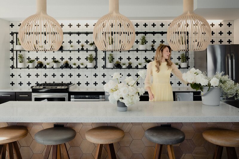 Woman in yellow dress and blonde hair is in large modern kitchen with graphic backsplash. She is working with a 2 buckets of white roses.