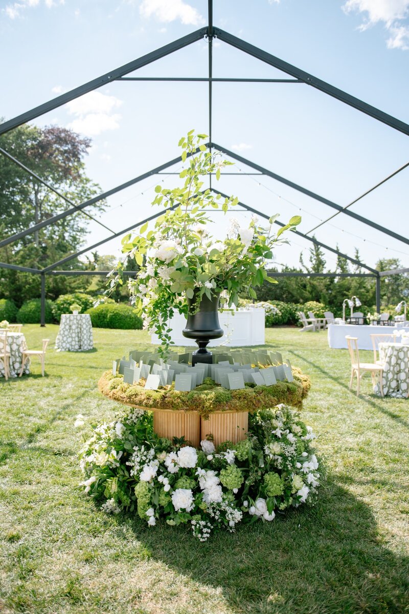 A circular escort card table surrounded by green and white flowers under a wedding tent