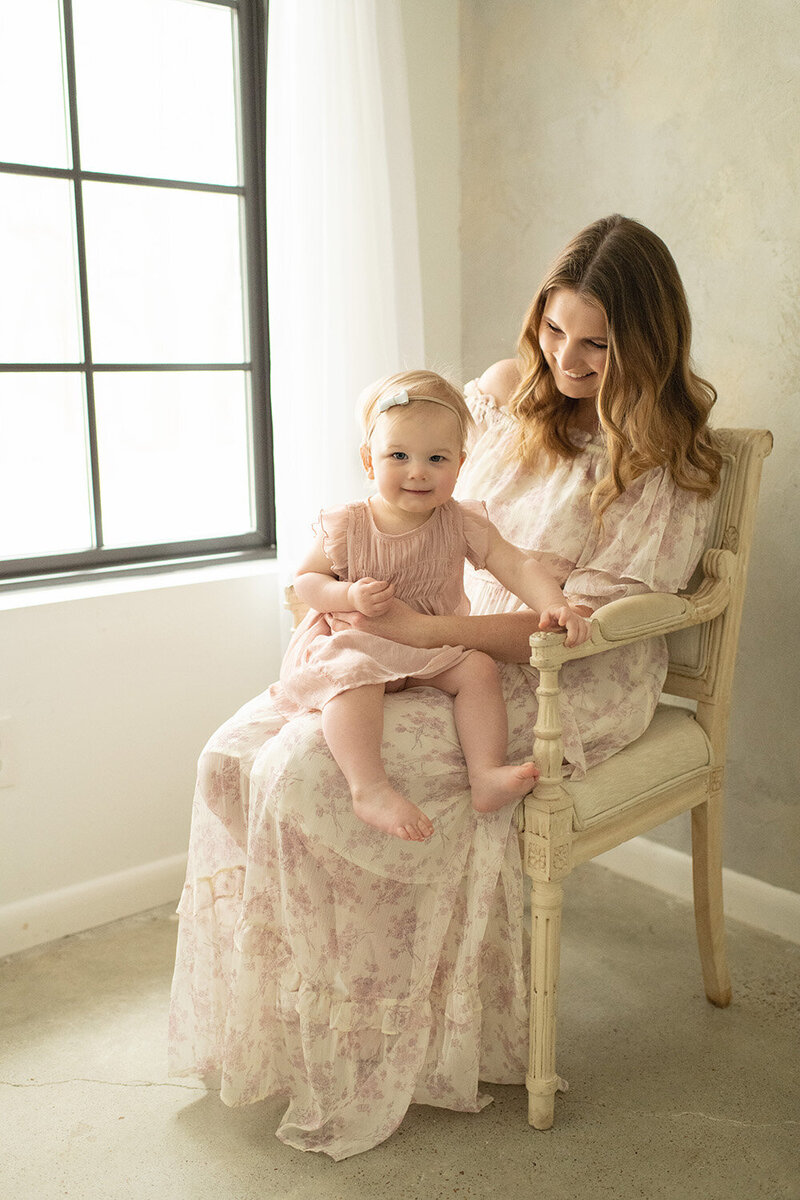 Mother in a floral dress sits on a vintage chair, holding her smiling toddler near a bright window. A heartfelt baby session by Moments in Light Photography.