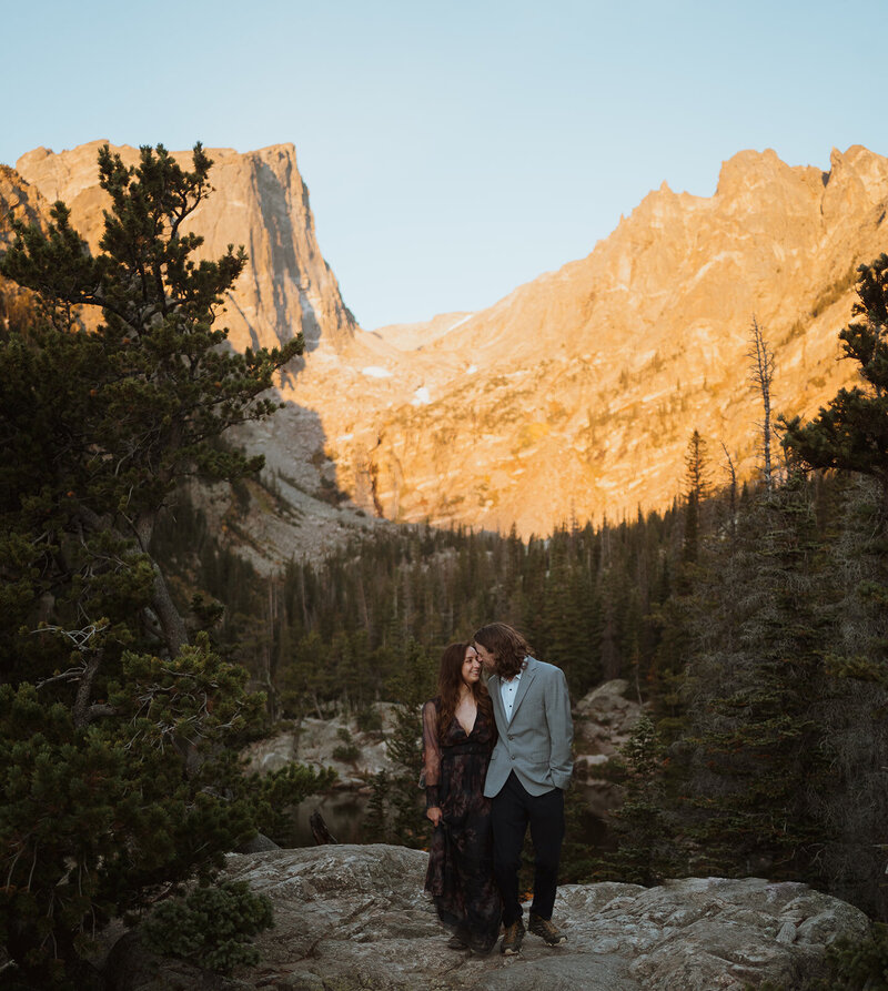 bride and groom are looking at each other and smiling. they are holding hands and walking towards the camera. they are on a large rock with a mountain behind them that is bright orange from the sun