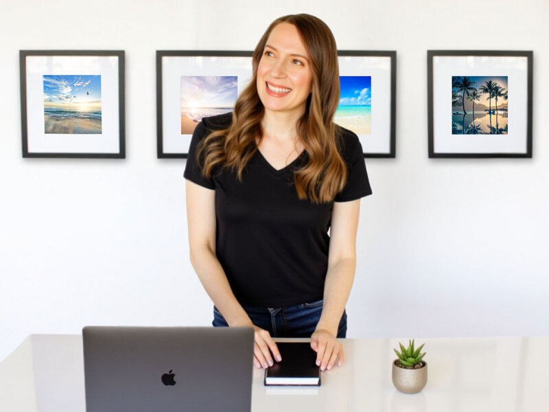 Stacey from Xanthe Bookkeeping smiling in a black V-neck t-shirt, standing behind a desk with a MacBook Pro and framed beach photos on the wall.