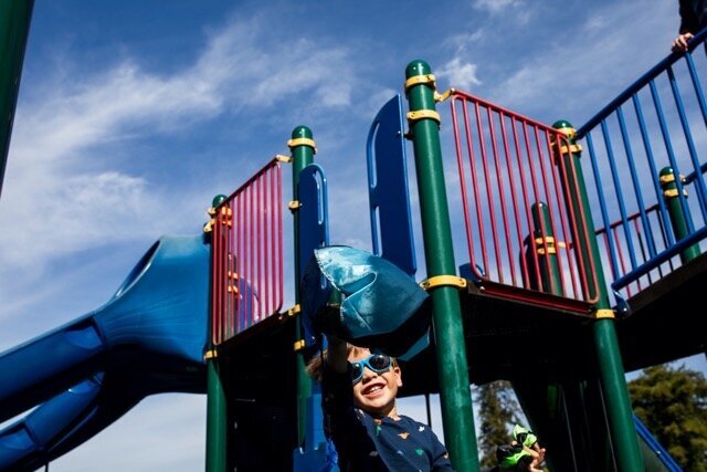 Little boy wearing blue sunglasses and a huge grin on his face reaches for a GI joe toy as it parachutes in front of him on the playground with slides in the background. 