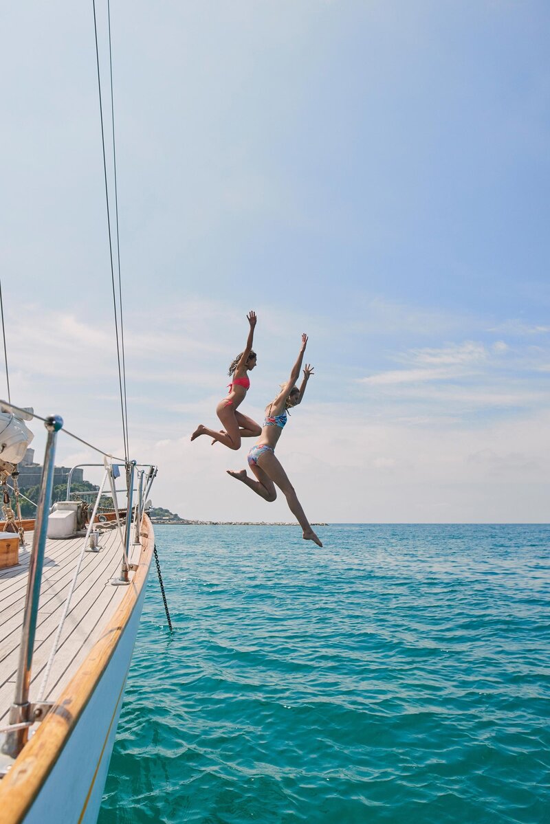 Two people in swimsuits are mid-air, joyfully jumping off a sailboat into the clear blue sea. The sailboat is on the left, with a sunny sky and calm water surrounding them.