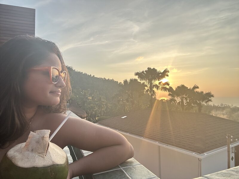 Maya is wearing orange rimmed sunglasses and has a coconut in her hand. She looks off into the sunset