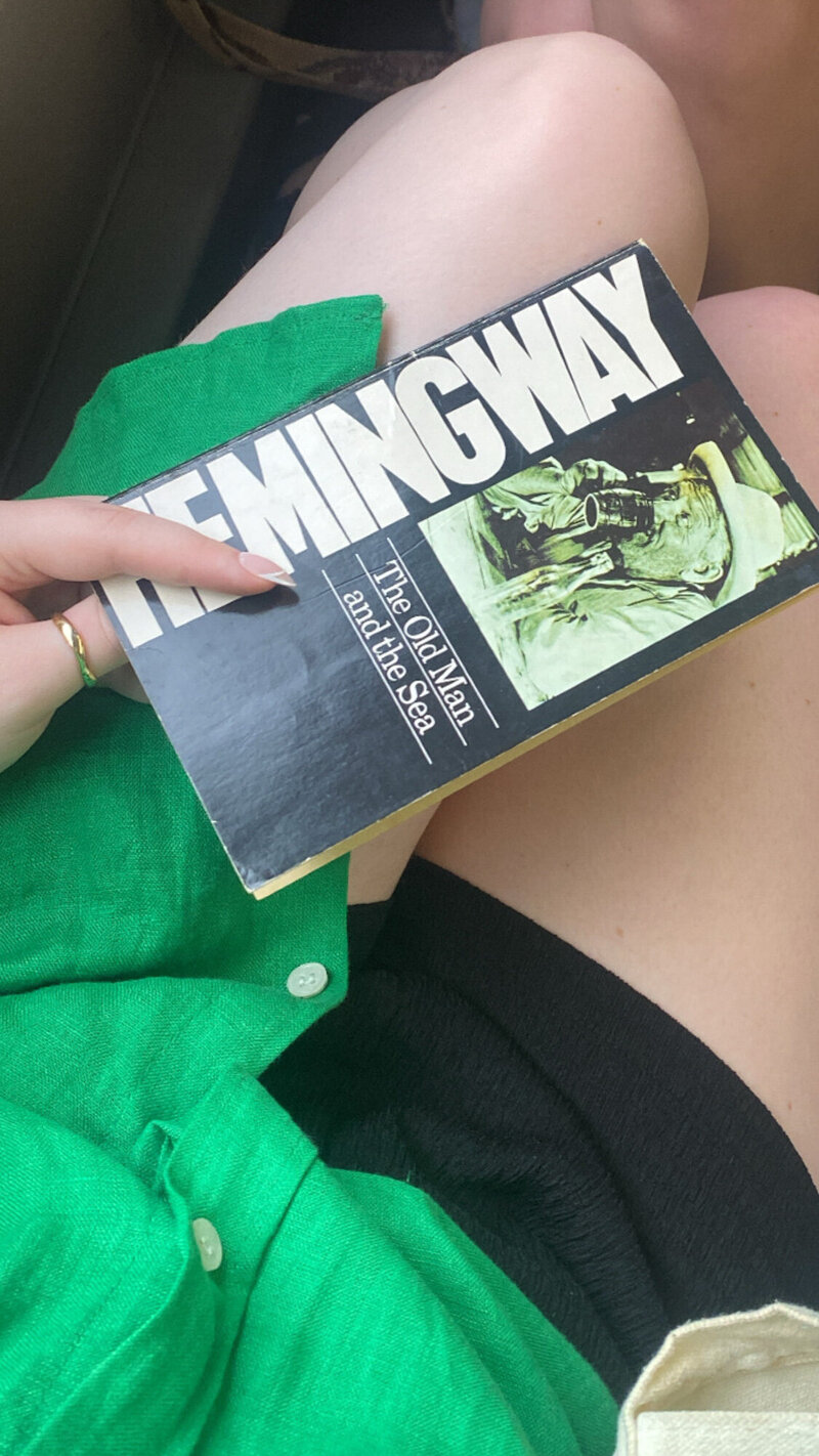 Person holding a copy of "The Old Man and the Sea" by Ernest Hemingway, wearing a green shirt and black skirt