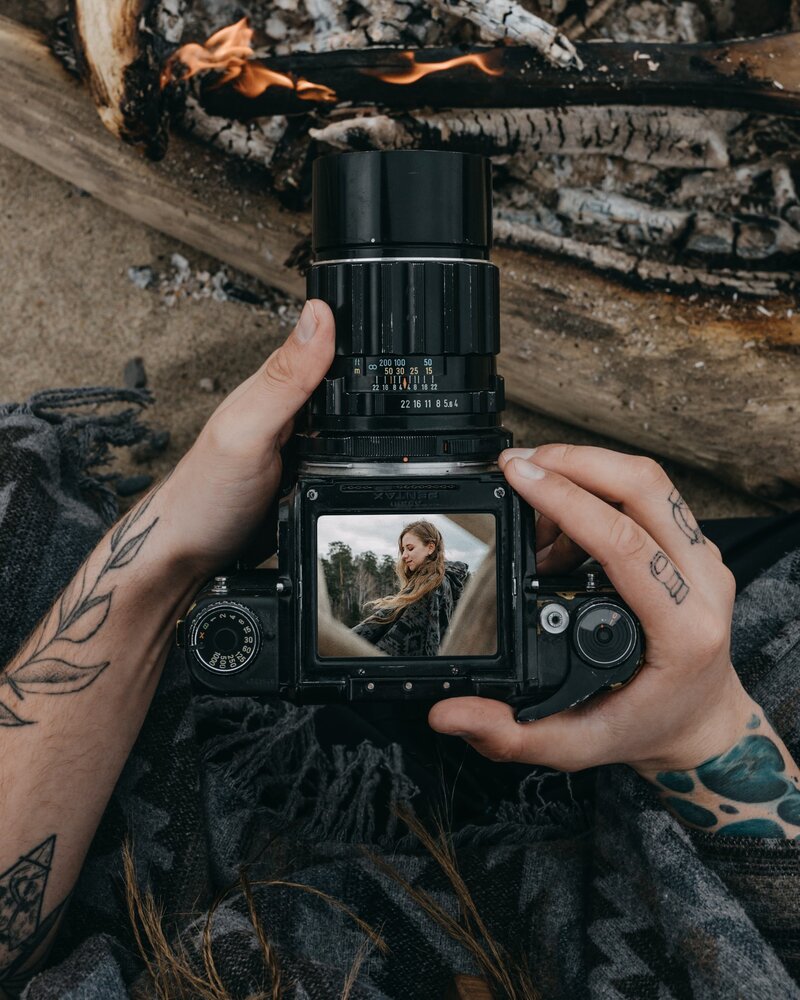 Man with tattooed arms holding a camera over a fire; camera is displaying a picture of a woman by a forest