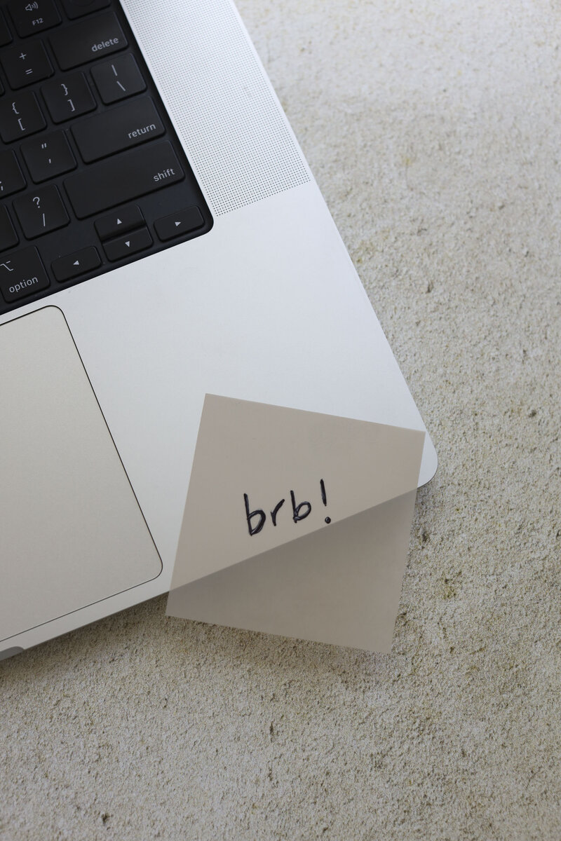 Close-up of a laptop keyboard with a sticky note that reads “brb!” on a neutral stone surface.