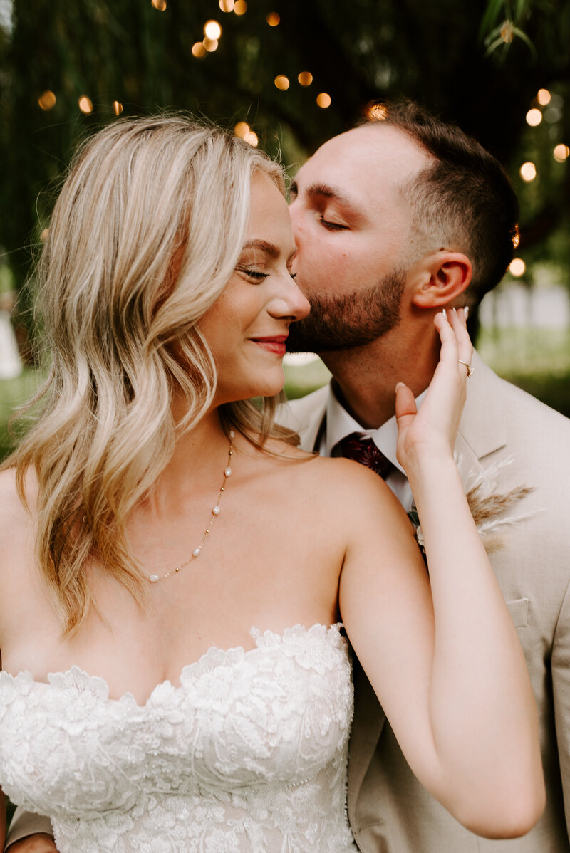 A groom kissing his bride's cheek while she closes her eyes and holds her bouquet.