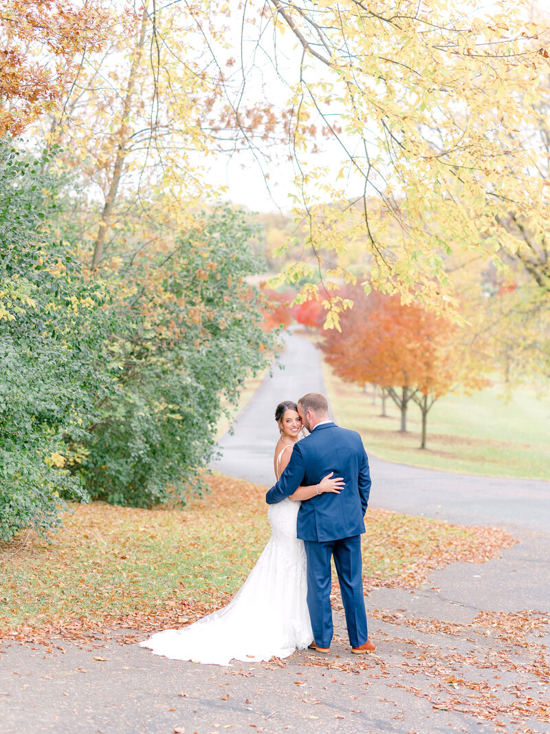 Bride and groom standing with their arms around each other outside under fall trees