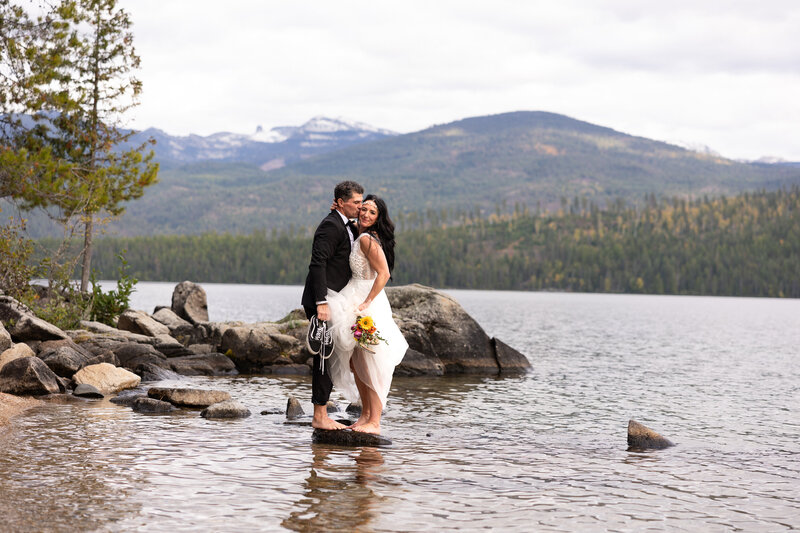Groom in tux bride in wedding dress holding flowers standing barefoot on rock in  Priest lake Idaho