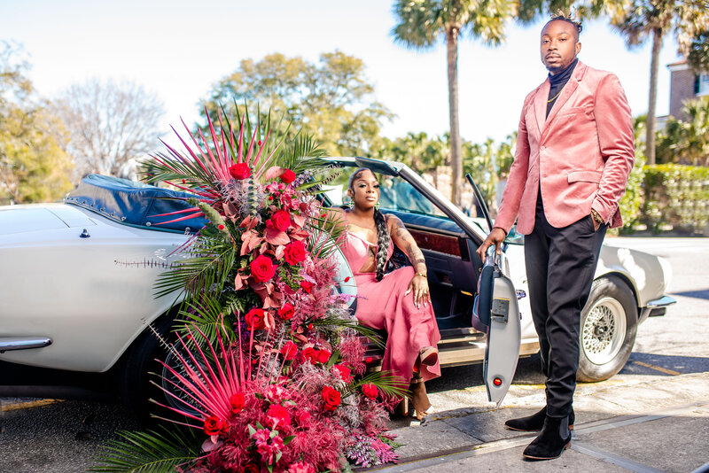 Capture the romance of a stunning Black couple in Charleston, framed by a vintage Aston Martin provided by Whipdashadorned with pink and white florals by Popout Decor. The woman, in a flowing pink gown from Fashion Nova, exudes elegance while her partner, in a stylish pink velvet blazer, creates a perfect moment of love and luxury.