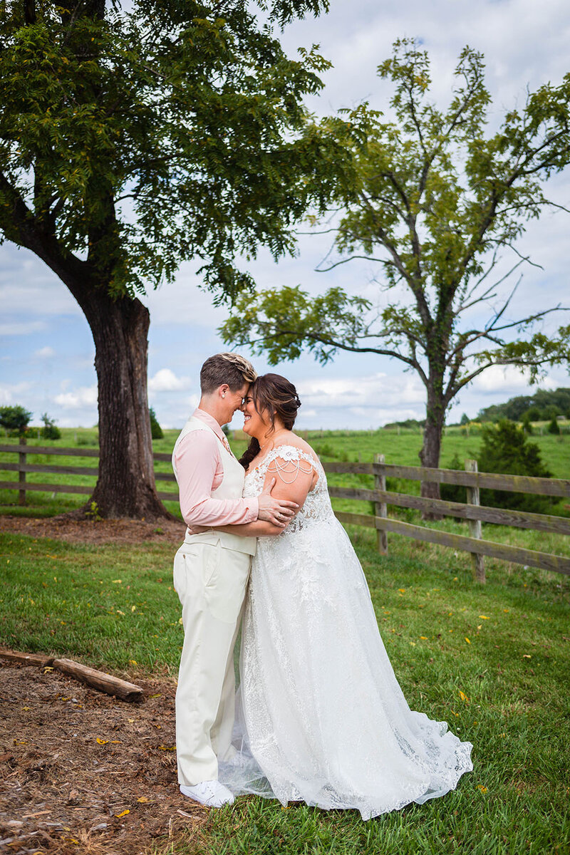 An LGBTQ+ couple embraces, touch foreheads, and smile at one another on their wedding day in Union Hall, Virginia in a sprawling landscape at the Pavilion at Black Water Junction.