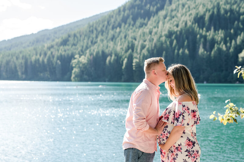 Man kissing pregnant woman's forehead by a lake