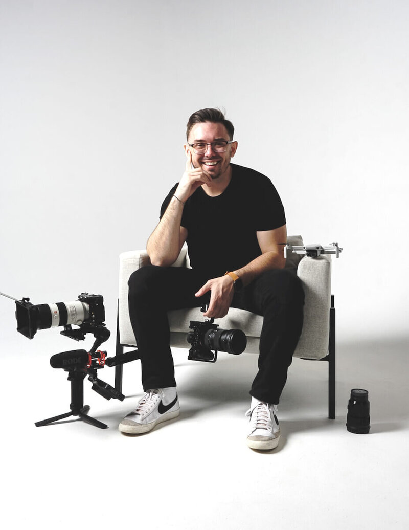 Founder of Atkinson Media House seated on a light gray chair, confidently holding a camera, amidst a backdrop of videographer equipment.