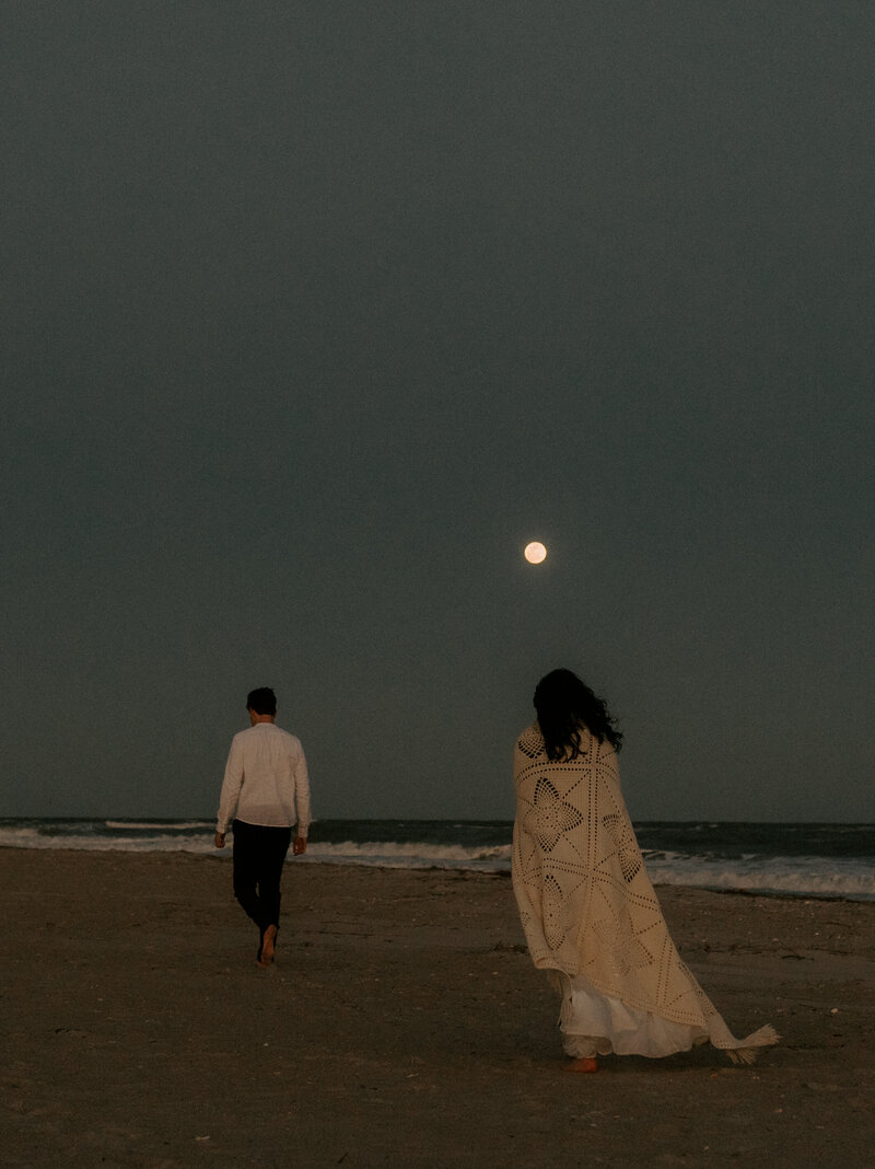 Married couple walking hand in hand along the beach under the moonlight, with soft waves glowing in the night.