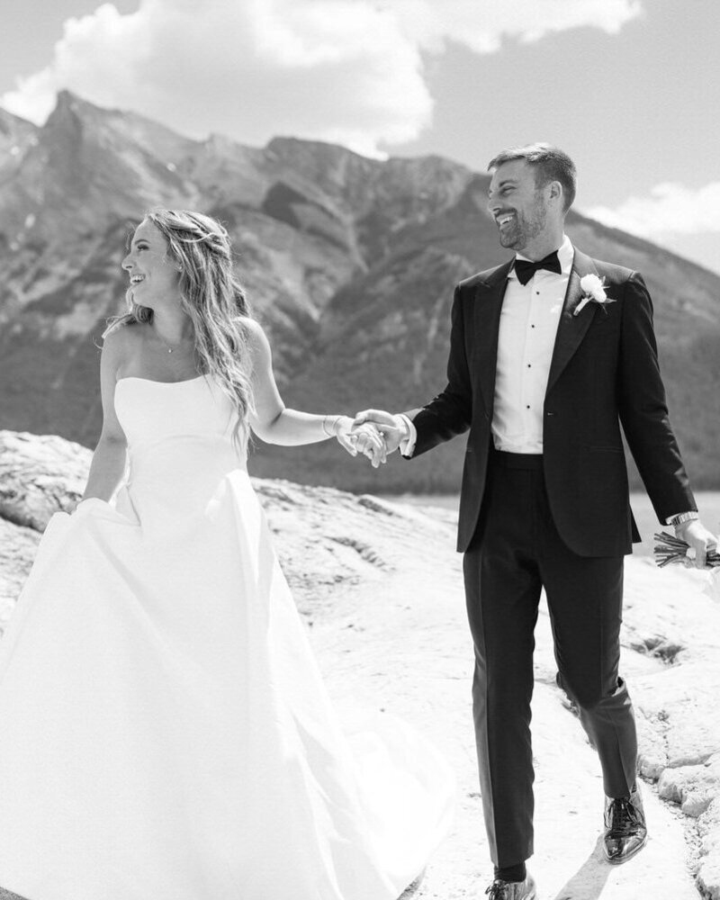 This joyful photograph captures the bride and groom walking hand in hand over rocks at Lake Minnewanka in Banff National Park. As they laugh together, the stunning natural beauty of the lake and mountains creates a perfect backdrop for their fun and intimate moment.