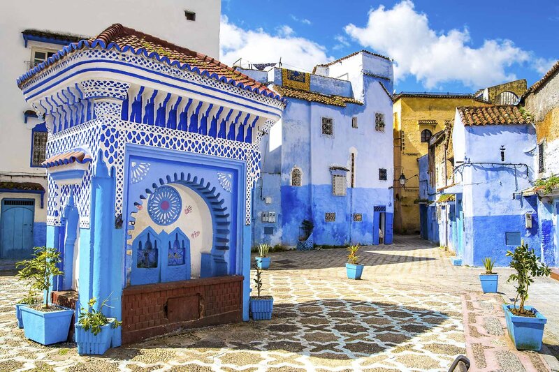 Colorful blue-washed buildings and ornate fountain in a sunny courtyard in Chefchaouen, Morocco.