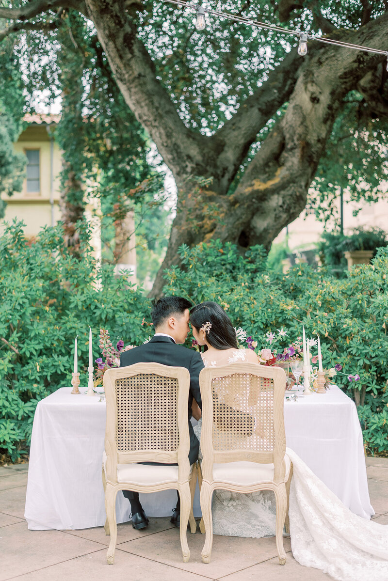 11-alisonbrynn-Radiant-LoveEvents-Maxwell-1-House-back-of-bride-groom-sitting-in-sweetheart-table-kissing-outdoors-bush-big-tree-backdrop-romantic-elegant-timeless