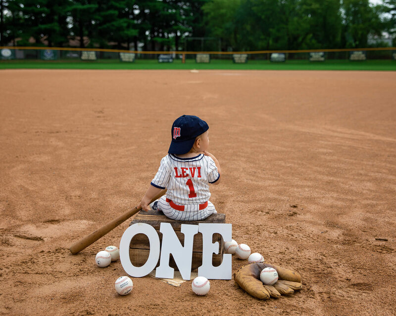 One year old boy sitting on a crate in a ball field. Surrounding him is a bat, balls, glove and the letters spelling out ONE.