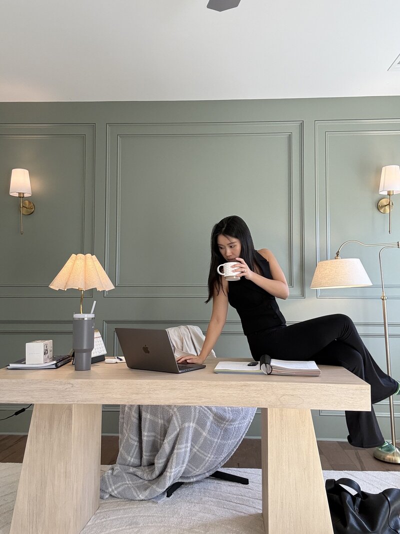 Stephy Chen sitting on a table with her laptop, creating content in a stylish green-paneled studio with warm lighting.