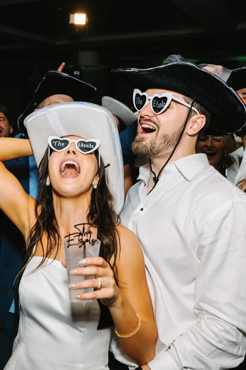 Bride and groom wearing cowboy hats and heart sunglasses while dancing