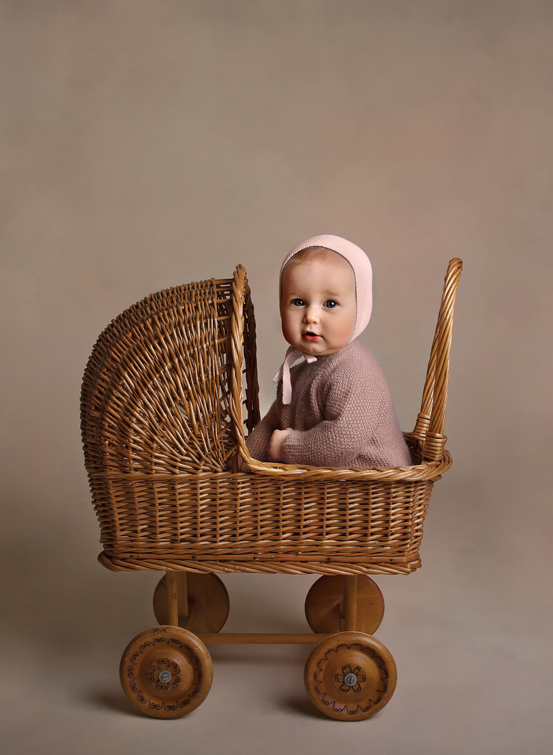 Brooklyn first birthday photoshoot. Baby girl in a dusty rose cardigan and bonnet is sitting in a miniature wicker pram. She is looking over her shoulder and smiling at the camera.  Captured by best Brooklyn, NY first birthday photographer Chaya Bornstein.