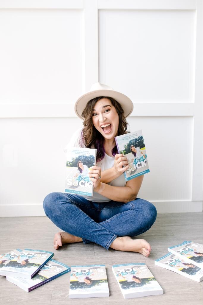 Melyssa Allen author holds up copies of her book Healthy Living Doesn't Have to Suck while she sits cross legged on the floor surrounded by more copies of her book. 