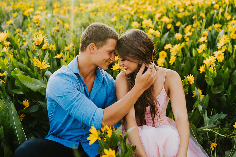 Couples photographer located in Rexburg Idaho, Adriana Montoya Photo. A photo of a couple sitting in a field of yellow flowers. He has his hand  going across his body and playing gently with her hair. Their foreheads and touching, both are smiling.