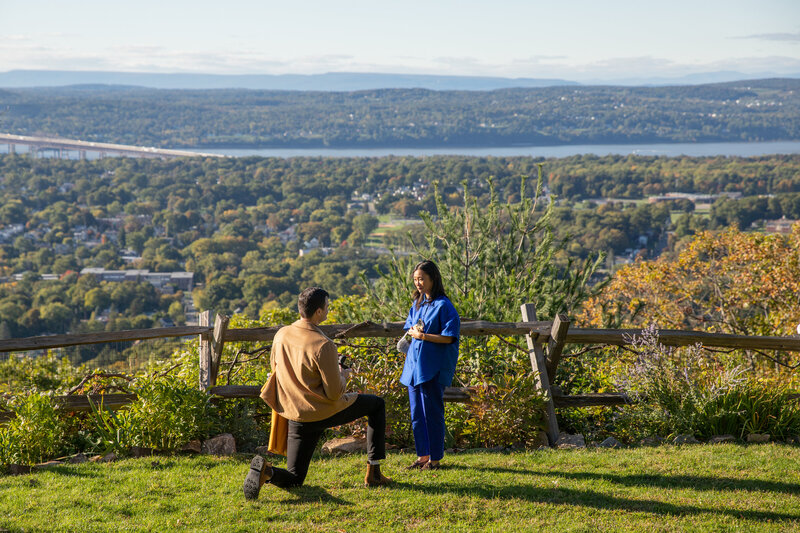 man kneels on one knee proposing to girlfriend with Hudson River and mountains in background in Beacon, NY