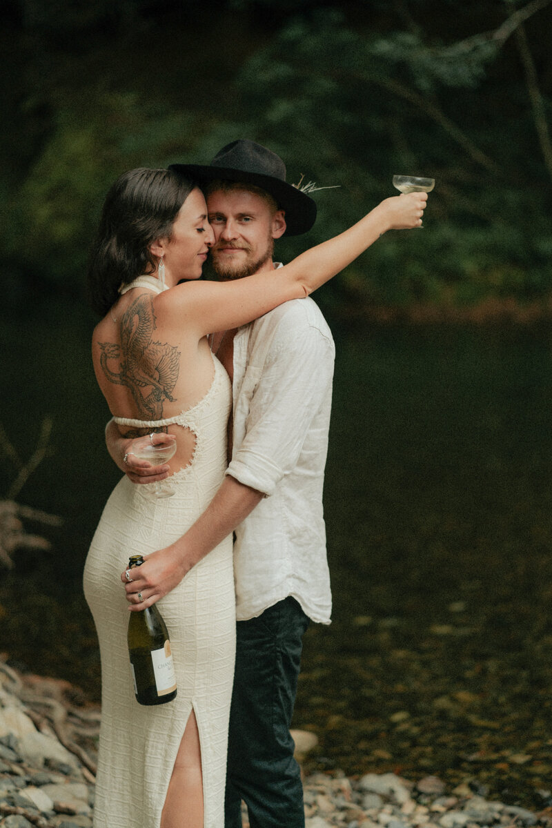 A rock and roll photo of a couple holding champagne on a riverbank  during their steamy couples shoot in Bellingen by Coffs Harbour photographer Fire & Fly