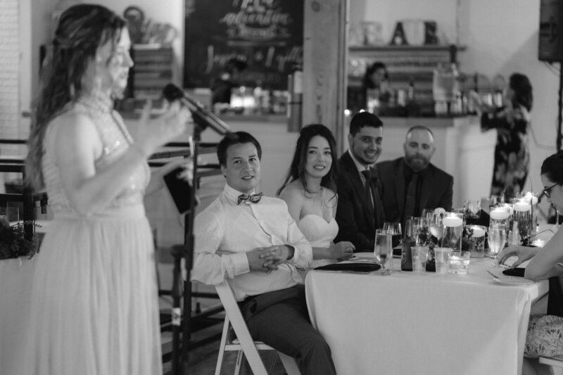 A woman delivers a speech at an event as guests seated at a table listen attentively.