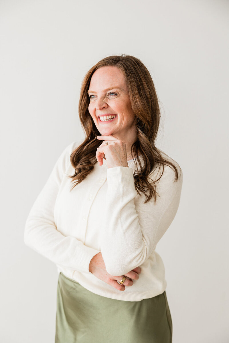 Woman in white sweater with red hair and curls, smiling off into a window
