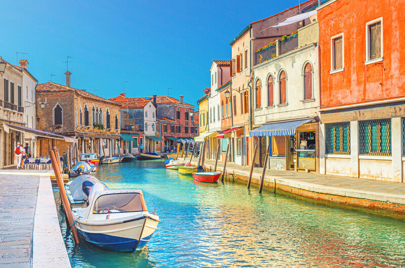 Colorful canal scene on Murano Island near Venice, Italy, with small boats and pastel buildings—part of the Venetian day trip experience on the Wish You WERE Here Italy Tour with author Christy Schillig.