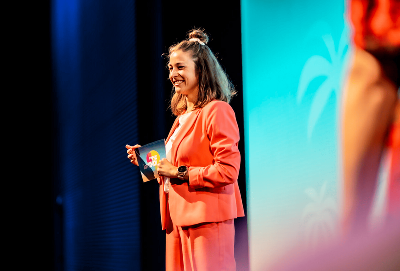 Eventmoderatorin Hanna Zöller auf einer Konferenz, lächelnd mit Moderationskarte auf der Bühne im roten Anzug