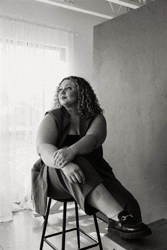 A black-and-white image of Andrea Cakmar sitting thoughtfully on a tall stool, dressed in a tan suit, looking away from the camera towards a light-filled window.